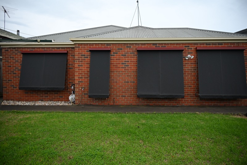 Four charcoal auto spring awnings fully extended across the exterior windows of a red brick home, viewed from the backyard