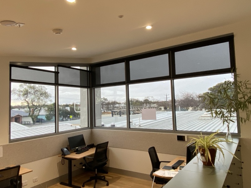 Charcoal Solarcool roller blinds fitted across wrap-around corner windows in a commercial office, reducing glare while preserving the rooftop view